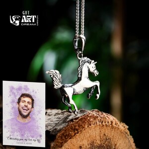Puede incluir: Un colgante de unicornio plateado suspendido de una cadena. El unicornio est&aacute; representado en posici&oacute;n encabritada. La imagen tambi&eacute;n incluye una tarjeta con una foto y el texto "Celebrating you, my love, my life" y los nombres Olivia & Adam.