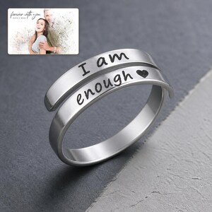 K&ouml;nnte beinhalten: Ein silberfarbener Wickelring mit der eingravierten Aufschrift I am enough und einem kleinen Herzsymbol. Der Ring hat eine glatte, polierte Oberfl&auml;che und ein offenes Design. Ein kleines Bild zeigt ein Paar mit dem Text forever with you Olivia & Adam.