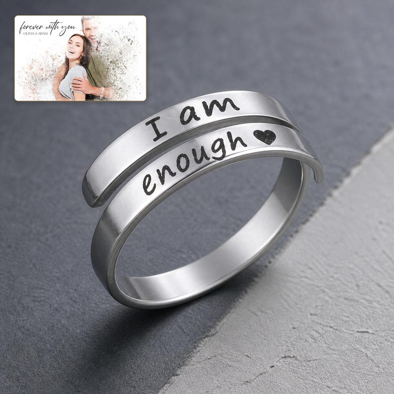 K&ouml;nnte beinhalten: Ein silberfarbener Wickelring mit der eingravierten Aufschrift I am enough und einem kleinen Herzsymbol. Der Ring hat eine glatte, polierte Oberfl&auml;che und ein offenes Design. Ein kleines Bild zeigt ein Paar mit dem Text forever with you Olivia & Adam.