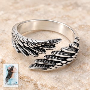 May include: A silver-toned open ring featuring a detailed wing design with textured feathers. The band is adjustable, with the two ends of the wing overlapping slightly. The metal has a polished finish with darkened recessed areas to highlight the feather details.