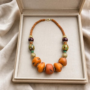 Puede incluir: Un collar de cuentas con peque&ntilde;as cuentas de madera naranja, piedras naranjas lisas m&aacute;s grandes, esferas de color granate oscuro y cuentas de vidrio multicolor con patrones. Incluye espaciadores de metal dorado y un cierre de mosquet&oacute;n.