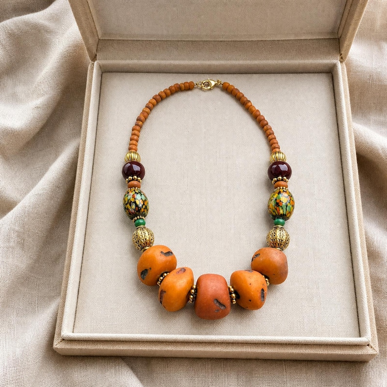 Puede incluir: Un collar de cuentas con peque&ntilde;as cuentas de madera naranja, piedras naranjas lisas m&aacute;s grandes, esferas de color granate oscuro y cuentas de vidrio multicolor con patrones. Incluye espaciadores de metal dorado y un cierre de mosquet&oacute;n.