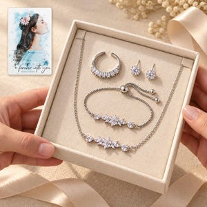 Two adults passing an open ivory jewelry box containing the floral necklace, matching bracelet, baguette ring and round stud earrings. A personalized digital greeting card is shown alongside.