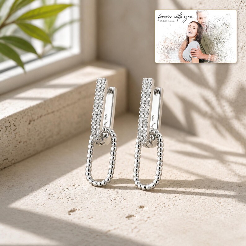May include: A pair of silver-toned drop earrings featuring a rectangular post encrusted with small clear stones and a dangling oval link made of beaded metal. A small rectangular card displays a photo of a couple with the text forever with you.