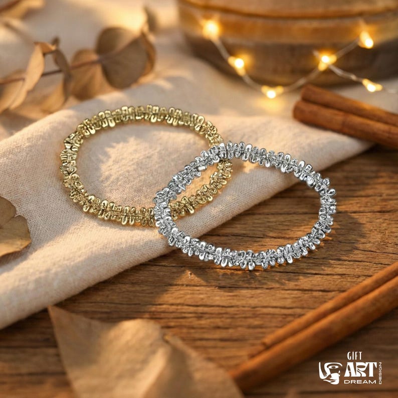 May include: Two decorative bracelets, one gold and one silver, rest on a cream-colored cloth. The bracelets have a textured, beaded design. Cinnamon sticks and a string of fairy lights are in the background, on a wooden surface.