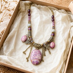 May include: A necklace featuring a large oval purple and white speckled stone pendant, accented with smaller matching oval beads, round purple and light gray beads, ornate antique brass-toned metal spacers, and dangling brass-toned chains.