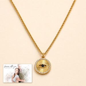 Gold-tone necklace with a round sunburst and black eye detail, display view. A personalized greeting-card example appears in the corner.