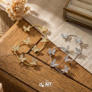 May include: Two ear cuffs, one gold and one silver, each decorated with multiple butterfly-shaped details. The butterflies are encrusted with small, sparkling crystals. The cuffs are displayed on a wooden surface.