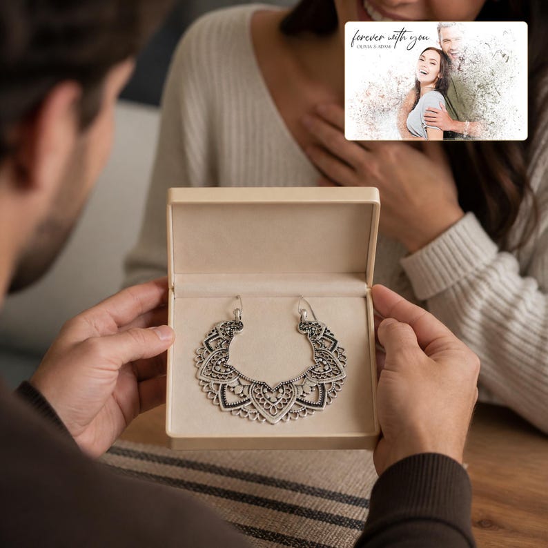 K&ouml;nnte beinhalten: Eine silberfarbene Halskette mit einem komplizierten, halbkreisf&ouml;rmigen Filigrananh&auml;nger mit herzf&ouml;rmigen Motiven, pr&auml;sentiert in einer beigen rechteckigen Geschenkbox. Eine kleine Karte mit einem Foto eines Paares und dem Text "forever with you" ist sichtbar.