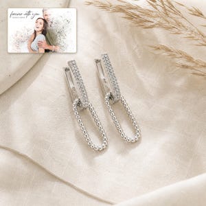 May include: A pair of silver-toned hoop earrings featuring a rectangular bar encrusted with small clear stones and a dangling oval link chain made of tiny metallic beads. A small rectangular card displays a photo of a couple with the text forever with you.