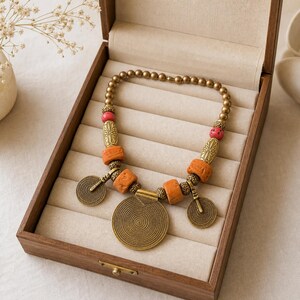 May include: A necklace featuring a series of small metallic beads, interspersed with textured orange cylindrical beads, ornate gold-toned metal spacers, and small red beads. Three circular metal pendants with spiral patterns hang from the center.