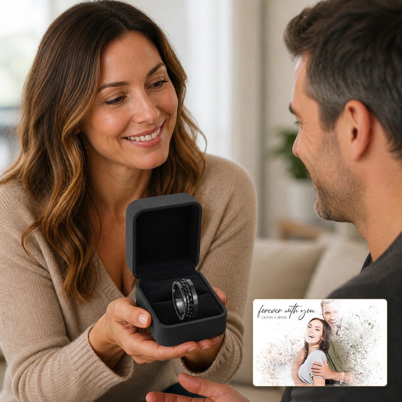 May include: A black ring with a central spinning chain detail, presented inside a square black velvet-lined jewellery box. Beside the box is a rectangular card featuring a printed photograph of a couple and the text "forever with you OLIVA & ADAM".