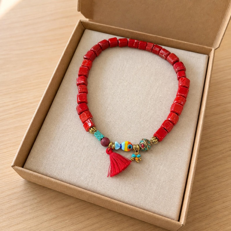 Op de afbeelding: Een armband met vierkante rode kralen, een klein rood kwastje en decoratieve bedels waaronder een veelkleurige vis, een donkerrode bol en goudkleurige accenten. De armband wordt gepresenteerd in een open kartonnen geschenkdoos.