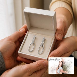 May include: A pair of silver-toned drop earrings featuring a rectangular top bar encrusted with small clear stones and a dangling oval link with a beaded texture, presented in a white jewellery box with a card reading forever with you.
