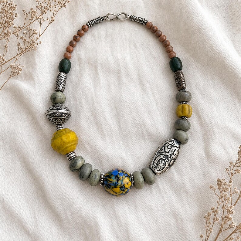 Puede incluir: Un collar de cuentas con una cuenta central redonda con patrones moteados en azul y amarillo, flanqueada por cuentas de piedra gris, espaciadores de metal plateado texturizado, una cuenta amarilla grande y cuentas de madera marr&oacute;n con cierre.