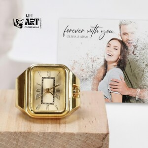 May include: A gold-toned watch ring with a rectangular face, Roman numeral hour markers, and black hands. The ring is displayed on a wooden block. The background includes a photo with the text "forever with you" and the names "Olivia & Adam".