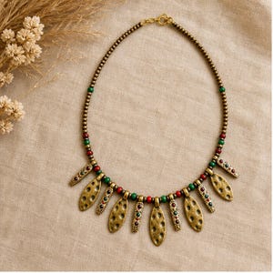 May include: A necklace featuring a series of small metallic beads in a bronze tone, interspersed with round red and green beads. The lower section includes several dangling, textured, oval-shaped metallic pendants with small inlaid red and green accents.