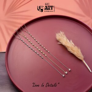 May include: Four delicate necklaces with small, sparkling stones are displayed on a maroon tray, alongside a dried ornamental grass. The necklaces vary in color, with silver and gold tones. The phrase "Love In Details" is written at the bottom.