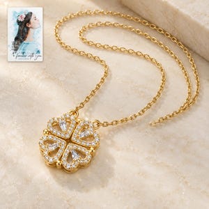 May include: A gold-tone necklace featuring a pendant shaped like a four-leaf clover, adorned with small clear stones and baguette-cut crystals. The pendant is designed to fold into a heart shape. A small card with a watercolor illustration and the text "forever with you" is visible.
