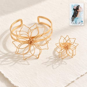 Puede incluir: Un brazalete y un anillo a juego de metal en tono dorado, ambos con un intrincado diseño floral de alambre abierto. Se incluye una pequeña tarjeta rectangular que muestra una ilustración y el texto "forever with you".