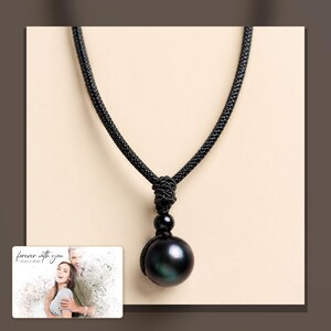 Black cord necklace with a dark round pendant, alternate framed view. A personalized greeting-card example appears in the corner.