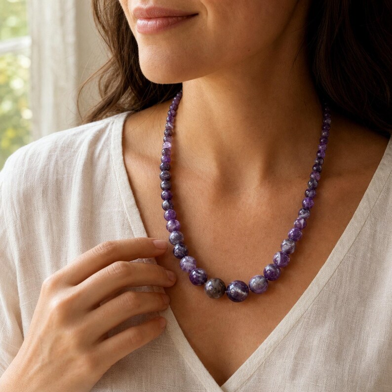 May include: A necklace featuring a strand of polished, spherical amethyst beads in varying shades of deep purple and violet. The beads are graduated in size, with the largest stones positioned at the centre and smaller beads tapering towards the ends.