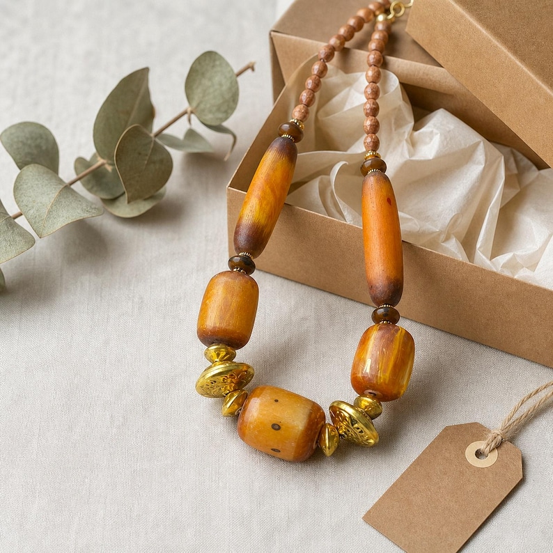 May include: A necklace featuring large, smooth, amber-colored cylindrical and barrel-shaped beads, interspersed with smaller dark brown spherical beads and textured gold-toned metal spacers, arranged on a string inside a brown cardboard gift box.