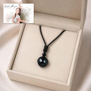 Black cord necklace with a dark round pendant, presentation in an open jewelry box. A personalized greeting-card example appears in the corner.