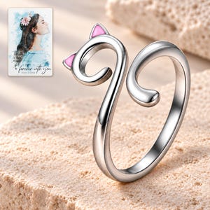 Complete polished silver-tone cat-curl ring on pale limestone, with two pink ear insets and an open tail gap. A personalized digital greeting card is shown alongside.