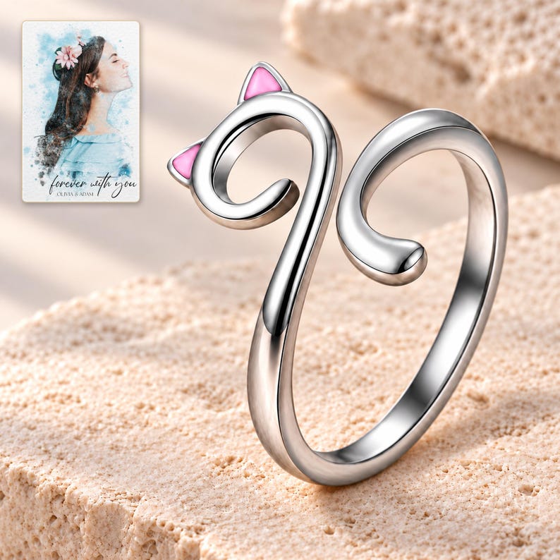 Complete polished silver-tone cat-curl ring on pale limestone, with two pink ear insets and an open tail gap. A personalized digital greeting card is shown alongside.
