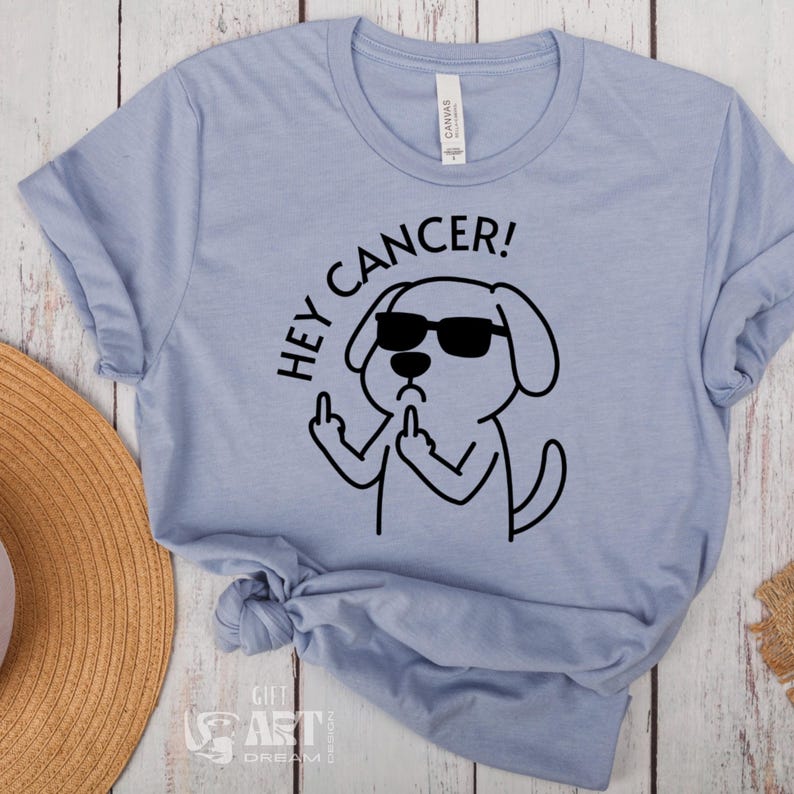 May include: A light blue t-shirt with a black graphic of a dog wearing sunglasses and giving the middle finger. The text "Hey Cancer!" is written above the dog.