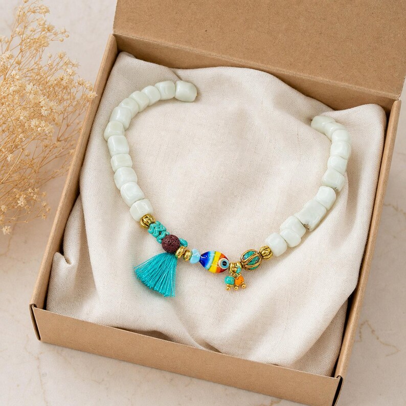 Puede incluir: Una pulsera de cuentas con cuentas cil&iacute;ndricas de color verde p&aacute;lido liso, acentuada con una peque&ntilde;a borla turquesa, una cuenta texturizada de color marr&oacute;n oscuro, un dije multicolor en forma de ojo y espaciadores de metal dorado, presentada en una caja.