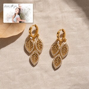 May include: A pair of gold hoop earrings featuring three dangling leaf-shaped charms encrusted with small clear stones. A rectangular card is visible in the corner with the text "forever with you" and a photograph of two people.