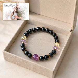 May include: A beaded bracelet featuring smooth black spherical beads, faceted yellow and purple translucent beads, and small metallic gold-toned spacers. A small rectangular card displays a photo of a couple with the text forever with you.