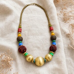 Op de afbeelding: Een kralenketting met een mix van materialen, waaronder grote bollen van geborsteld goudkleurig metaal, getextureerde donkerbruine ronde kralen, levendige rode, blauwe, groene en gele cilindrische en bolvormige kralen en kleine goudkleurige tussenkralen.