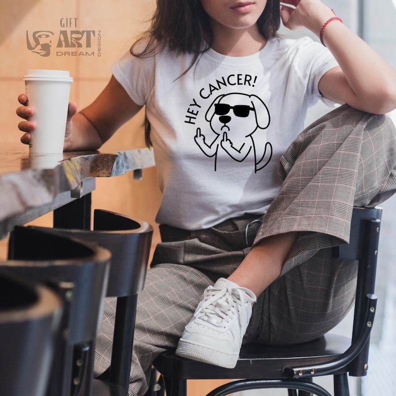May include: White t-shirt with a black and white illustration of a dog wearing sunglasses and giving the middle finger. The text "Hey Cancer!" is above the dog.