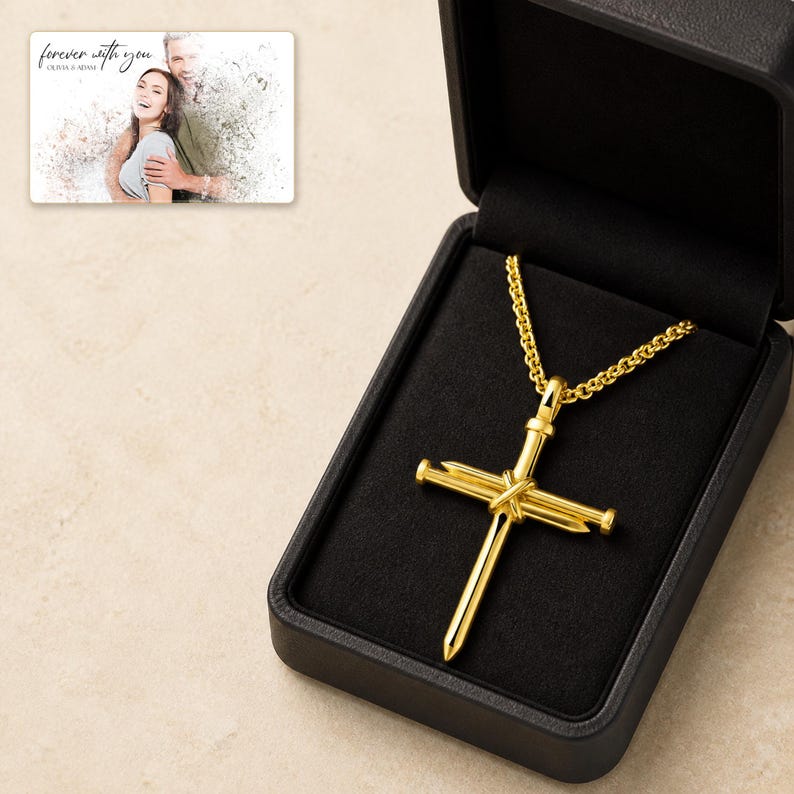 K&ouml;nnte beinhalten: Eine goldfarbene Kreuz-Halskette mit einem Design aus zwei gekreuzten, zusammengebundenen N&auml;geln, die an einer passenden Kette h&auml;ngt und in einer mit schwarzem Samt ausgekleideten Schmuckschatulle pr&auml;sentiert wird, inklusive einer Fotokarte.