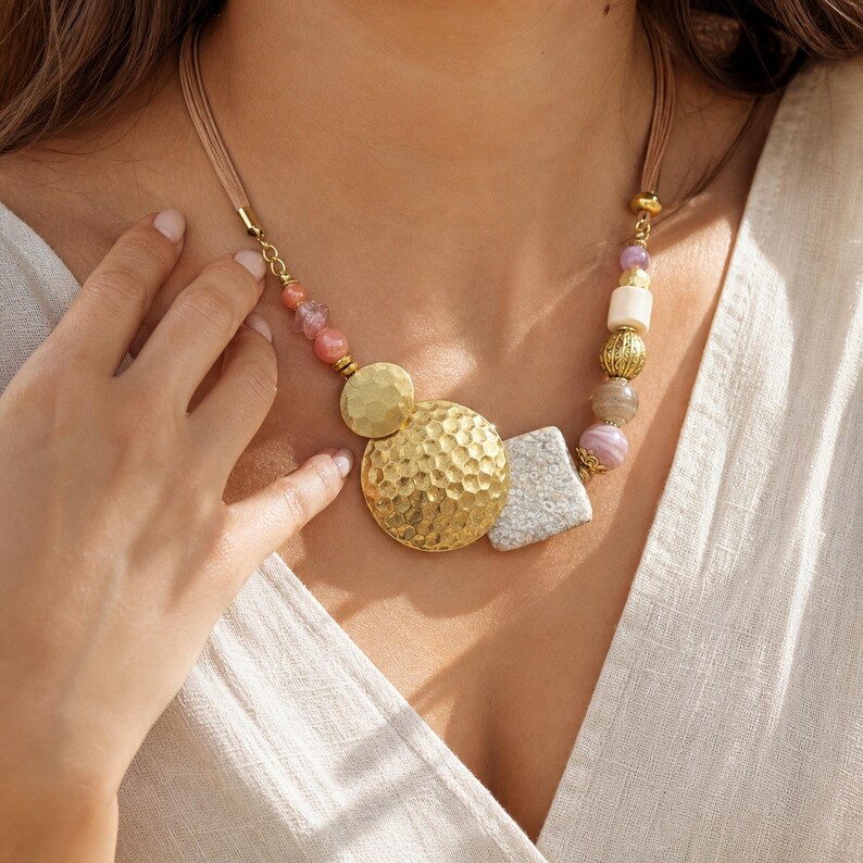 May include: A multi-strand cord necklace featuring two large, hammered gold-tone circular pendants, a textured grey stone-like piece, and an assortment of beads in shades of pink, white, and gold, arranged in a decorative, asymmetrical pattern.
