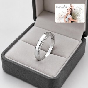 May include: A silver-toned metal ring with a textured, beaded edge detail resting inside a dark gray jewelry box. The box lid features a printed photograph of a couple with the text "forever with you" and "OLIVIA & ADAM" in script.