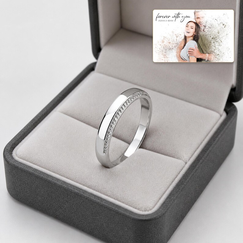 May include: A silver-toned metal ring with a textured, beaded edge detail resting inside a dark gray jewelry box. The box lid features a printed photograph of a couple with the text "forever with you" and "OLIVIA & ADAM" in script.