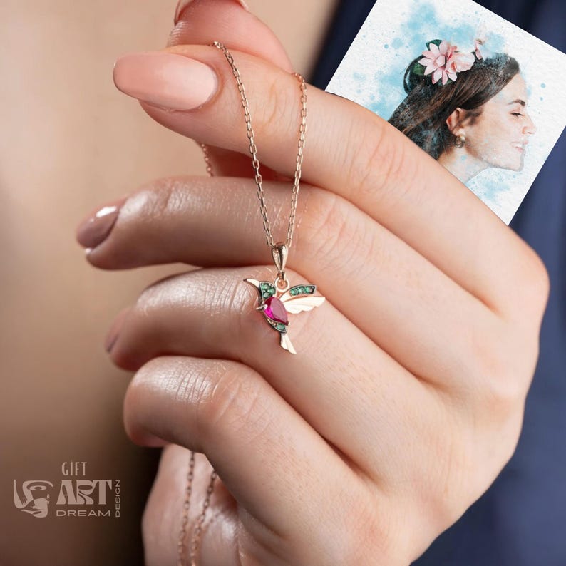 May include: A gold-colored hummingbird pendant necklace. The pendant features a ruby-colored gemstone body, green gemstone accents, and white wings. The necklace is held in a hand, with a watercolor portrait in the background. The logo "GIFT ART DREAM DESIGN" is visible.