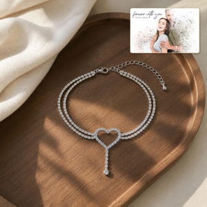 May include: A double-strand silver-toned anklet featuring a central heart-shaped charm encrusted with clear stones and a dangling stone accent. The jewelry includes a lobster clasp and an adjustable chain. A small card displays the text "forever with you" and "OLIVIA & ADAM" above a photograph of a couple.