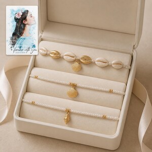 May include: A set of three bracelets in a cream-colored jewelry box. The top bracelet features white cowrie shells and a gold scallop charm. The middle and bottom bracelets consist of white seed beads with gold accents and gold shell charms.