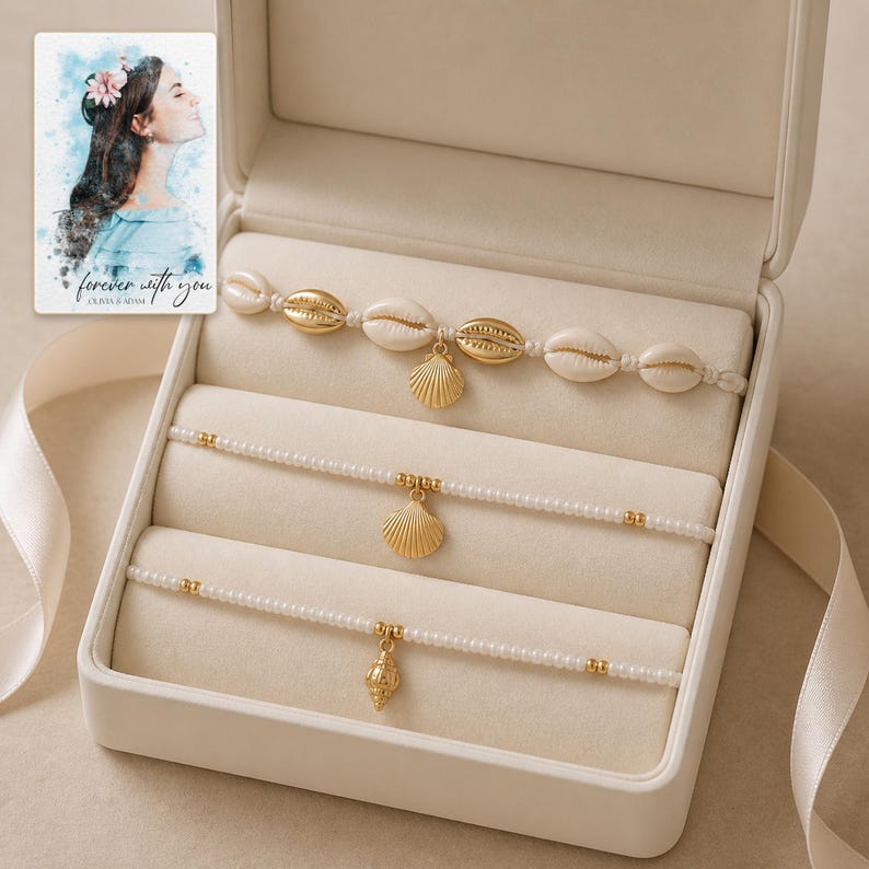 May include: A set of three bracelets in a cream-colored jewelry box. The top bracelet features white cowrie shells and a gold scallop charm. The middle and bottom bracelets consist of white seed beads with gold accents and gold shell charms.