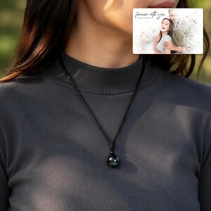Black cord necklace with a dark round pendant, worn view. A personalized greeting-card example appears in the corner.
