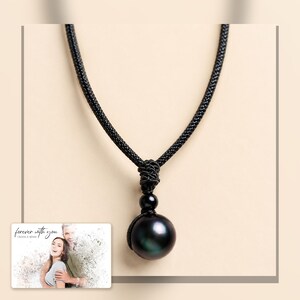Black cord necklace with a dark round pendant, framed product view. A personalized greeting-card example appears in the corner.