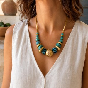 May include: A necklace featuring a central large gold-toned filigree bead flanked by alternating turquoise-colored disc beads and smaller gold-toned metallic beads, suspended from a chain of small gold-toned spherical beads on each side.