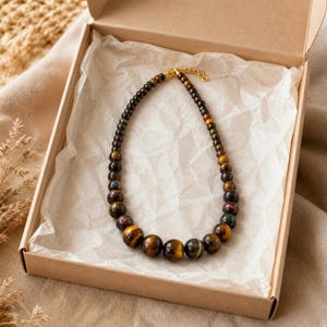 May include: A beaded necklace featuring polished tiger eye stones in varying shades of brown, gold, and dark grey. The beads increase in size towards the centre of the strand, which is finished with a gold-toned metal clasp and an adjustable chain.