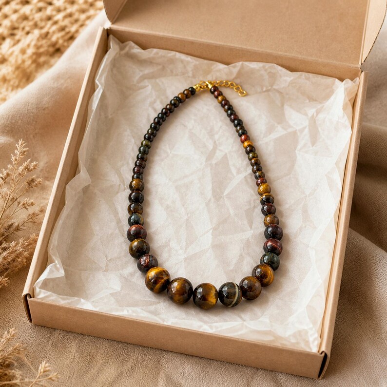 May include: A beaded necklace featuring polished tiger eye stones in varying shades of brown, gold, and dark grey. The beads increase in size towards the centre of the strand, which is finished with a gold-toned metal clasp and an adjustable chain.