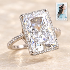 May include: A silver-toned ring featuring a large rectangular radiant-cut center stone surrounded by a halo of small round stones. The band is also encrusted with small round stones. A small card with a watercolor illustration and the text "forever with you" is visible.